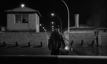 Movie still from “The Spy Who Came in from the Cold” (1965), directed by Martin Ritt – A man standing next to a bike on a street; Wide shot, Low angle