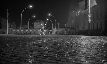 Movie still from “The Spy Who Came in from the Cold” (1965), directed by Martin Ritt – A person riding a bike on a paved road at night; Extreme Wide shot, High angle