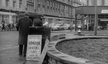 Movie still from “The Spy Who Came in from the Cold” (1965), directed by Martin Ritt – Two men standing in front of a newspaper stand in a city; Wide shot, Low angle