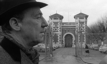 Movie still from “The Spy Who Came in from the Cold” (1965), directed by Martin Ritt – An older man wearing a hat standing in front of a gate; Close Up shot, Over the shoulder angle