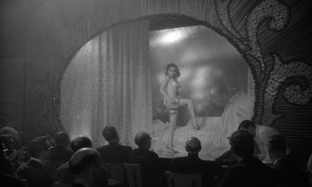 Movie still from “The Spy Who Came in from the Cold” (1965), directed by Martin Ritt – An old photo of people watching a woman in lingerie on a stage; Wide shot, Over the shoulder angle