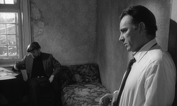 Movie still from “The Spy Who Came in from the Cold” (1965), directed by Martin Ritt – A man standing next to another man sitting on a couch; Medium shot, Over the shoulder angle
