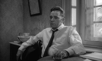 Movie still from “The Spy Who Came in from the Cold” (1965), directed by Martin Ritt – Black and white photograph of a man sitting at a table with a drink; Medium shot, Low angle