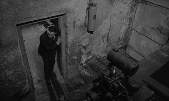 Movie still from “The Spy Who Came in from the Cold” (1965), directed by Martin Ritt – A man standing in a doorway next to an old machine; Wide shot, High angle