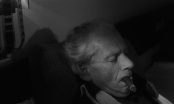 Movie still from “The State of Things” (1982), directed by Wim Wenders – An older man smoking a cigarette while sitting in a chair; Close Up shot, High angle