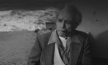 Movie still from “The State of Things” (1982), directed by Wim Wenders – An older man smoking a cigarette near the ocean; Medium shot, High angle