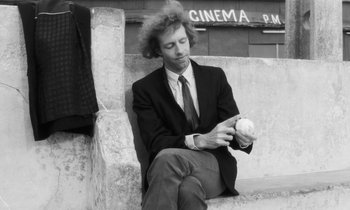 Movie still from “The State of Things” (1982), directed by Wim Wenders – A black and white photo of a man sitting on a bench holding a ball; Medium shot, Low angle