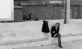 Movie still from “The State of Things” (1982), directed by Wim Wenders – A man sitting on the side of a building next to a sidewalk; Wide shot, Low angle