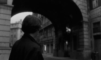 Movie still from “The State of Things” (1982), directed by Wim Wenders – A person standing on a sidewalk under an archway; Close Up shot, Over the shoulder angle