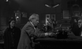 Movie still from “The State of Things” (1982), directed by Wim Wenders – An older man sitting at a bar with a woman sitting at the bar; Medium shot, Low angle