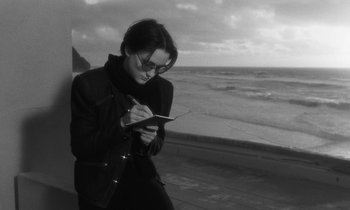 Movie still from “The State of Things” (1982), directed by Wim Wenders – A woman is writing on a notepad by the ocean; Medium shot, High angle