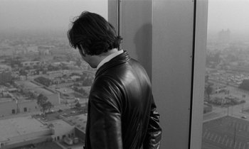 Movie still from “The State of Things” (1982), directed by Wim Wenders – A man in a leather jacket looking out a window at a city; Medium shot, Over the shoulder angle