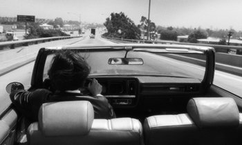 Movie still from “The State of Things” (1982), directed by Wim Wenders – A woman sitting in the driver's seat of a car on a highway; Wide shot, High angle