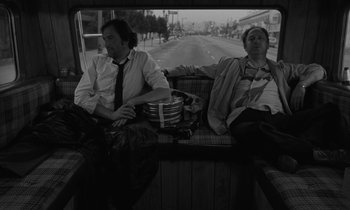 Movie still from “The State of Things” (1982), directed by Wim Wenders – Two men sitting on a bus with a road in the background; Medium shot, Low angle
