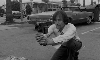 Movie still from “The State of Things” (1982), directed by Wim Wenders – A black and white photo of a man holding a video camera; Medium shot, Low angle