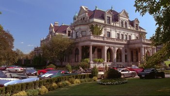 Movie still from “The Stepford Wives” (2004), directed by Frank Oz – A very big nice looking building with a lot of cars parked in front of it; Extreme Wide shot, Low angle