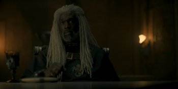 Movie still from “House of the Dragon” (2022), created by George R.R. Martin – An older man with dreadlocks sitting at a table; Medium shot, Over the shoulder angle