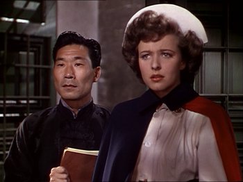 Movie still from “The Story of Dr. Wassell” (1944), directed by Cecil B. DeMille – An older man standing next to an older woman holding a book; Medium shot, Over the shoulder angle