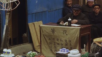 Movie still from “The Story of Qiu Ju” (1992), directed by Yimou Zhang – Two people sitting at a table with a sign on it; Wide shot, High angle