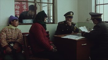 Movie still from “The Story of Qiu Ju” (1992), directed by Yimou Zhang – Two people sitting at a desk in a room; Medium shot, Over the shoulder angle