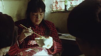 Movie still from “The Story of Qiu Ju” (1992), directed by Yimou Zhang – A woman in a red and black plaid shirt eats a bowl of noodles; Medium shot, High angle