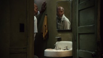 Movie still from “The Sunset Limited” (2011), directed by Tommy Lee Jones – A man standing in front of a bathroom mirror in front of a sink; Medium shot, Over the shoulder angle