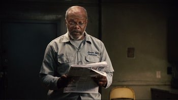 Movie still from “The Sunset Limited” (2011), directed by Tommy Lee Jones – An older man reading a newspaper in a room; Medium shot, Over the shoulder angle