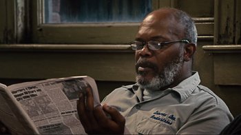 Movie still from “The Sunset Limited” (2011), directed by Tommy Lee Jones – An older man reading a newspaper while sitting in a chair; Close Up shot, Over the shoulder angle