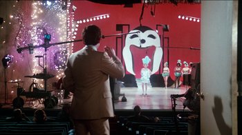 Movie still from “The Sunshine Boys” (1975), directed by Herbert Ross – A man standing in front of a stage with a large screen; Wide shot, Over the shoulder angle