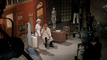 Movie still from “The Sunshine Boys” (1975), directed by Herbert Ross – A woman standing next to a man sitting on a chair; Wide shot, High angle