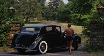 Movie still from “The Swimmer” (1968), directed by Sydney Pollack – A man in swim trunks standing next to an old car; Wide shot, High angle
