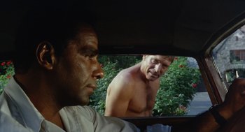 Movie still from “The Swimmer” (1968), directed by Sydney Pollack – A man looking in the mirror of a car while another man looks on; Close Up shot, Over the shoulder angle