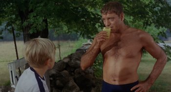 Movie still from “The Swimmer” (1968), directed by Sydney Pollack – A shirtless man drinking a beverage from a glass while standing next to a young boy; Medium shot, Over the shoulder angle