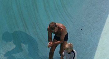 Movie still from “The Swimmer” (1968), directed by Sydney Pollack – A man standing next to a boy on a surfboard in the water; Wide shot, Overhead angle