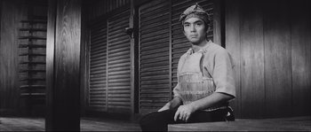 Movie still from “The Sword of Doom” (1966), directed by Kihachi Okamoto – A man sitting on top of a bench in front of a building; Medium shot, Low angle