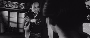 Movie still from “The Sword of Doom” (1966), directed by Kihachi Okamoto – A man in a black and white photo holding a donut; Medium shot, Over the shoulder angle