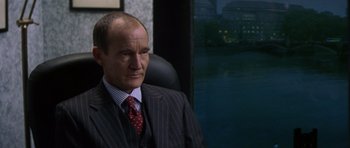 Movie still from “The Tailor of Panama” (2001), directed by John Boorman – A man in a suit sitting in a chair; Close Up shot, Over the shoulder angle