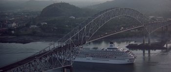 Movie still from “The Tailor of Panama” (2001), directed by John Boorman – A cruise ship in a body of water near a bridge; Extreme Wide shot, High angle
