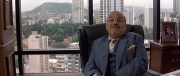Movie still from “The Tailor of Panama” (2001), directed by John Boorman – A man in a suit sitting in an office chair in front of a window; Close Up shot, High angle