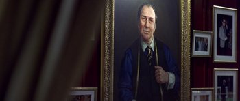 Movie still from “The Tailor of Panama” (2001), directed by John Boorman – An older man in a vest and tie is in front of a painting; Close Up shot, Low angle