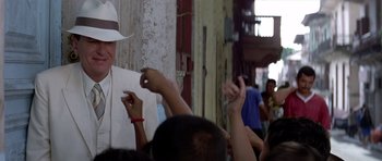 Movie still from “The Tailor of Panama” (2001), directed by John Boorman – A group of people gathered around a man in a white hat; Medium shot, Over the shoulder angle