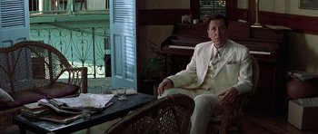 Movie still from “The Tailor of Panama” (2001), directed by John Boorman – A man in a white suit sitting on a chair; Medium shot, High angle