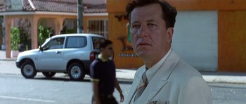 Movie still from “The Tailor of Panama” (2001), directed by John Boorman – A man standing on the side of the road with a car in the background; Close Up shot, Low angle