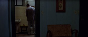 Movie still from “The Tailor of Panama” (2001), directed by John Boorman – A man standing in front of a sink in a room; Wide shot, Over the shoulder angle