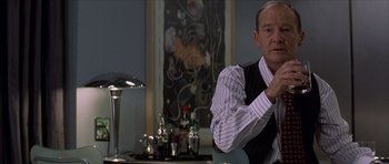 Movie still from “The Tailor of Panama” (2001), directed by John Boorman – A man sitting at a table with a glass of wine; Medium shot, Over the shoulder angle