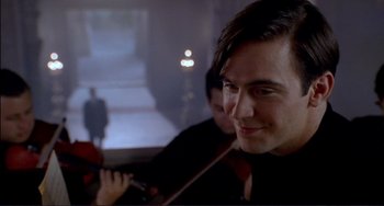 Movie still from “The Talented Mr. Ripley” (1999), directed by Anthony Minghella – A man smiles while holding a violin in front of another man; Close Up shot, Over the shoulder angle