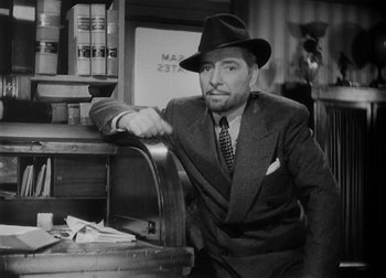 Movie still from “The Talk of the Town” (1942), directed by George Stevens – A man in a suit and hat leaning on a desk; Medium shot, Low angle