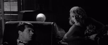 Movie still from “The Tarnished Angels” (1957), directed by Douglas Sirk – A black and white photo of a woman sitting on a couch next to a clock; Close Up shot, Over the shoulder angle