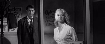 Movie still from “The Tarnished Angels” (1957), directed by Douglas Sirk – A man and a woman standing next to each other in front of an advertisement; Medium shot, Low angle