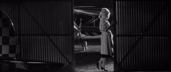 Movie still from “The Tarnished Angels” (1957), directed by Douglas Sirk – A black and white photo of a woman in a trench coat and high heels; Wide shot, Low angle